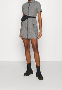 Musta-valkoinen ruutukuvioinen lyhyt mekko, jossa on etuvetoketju, lyhyet hihat ja kaksi taskua, yhdistettynä mustiin nyörikenkiin ja vyölaukkuun.