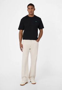 Musta lyhythihainen t-paita, jossa vihreä logo, yhdistetty vaaleanbeigeihin leveälahkeisiin housuihin ja valkoisiin kumipohjaisiin lenkkareihin.