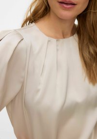 Beige satengbluse med rund hals, myke folder langs skuldrene og en glatt tekstur, som fremhever et minimalistisk design.