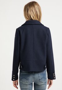 Žena ve tmavě modré zkrácené bundičce s knoflíkovými detaily na rukávech a zadní části, oblečená do světlých modrých džínů, stojící zády.