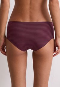 Eine Frau trägt lila und schwarze horizontal gestreifte Hipster-Unterw�äsche, gezeigt von der unteren Rückseite bis zu den oberen Oberschenkeln vor einem einfarbigen Hintergrund.