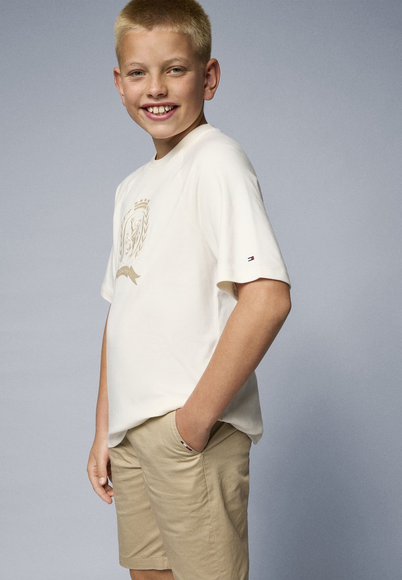 Kurzarm-Creme T-Shirt mit goldfarbigem Logo-Design auf der Brust; kombiniert mit beigen Shorts. Das Shirt hat einen kleinen Logo-Akzent auf dem Ärmel.