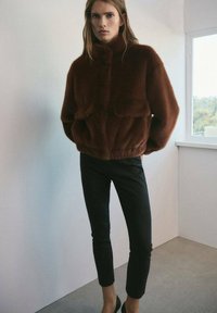 Donna che indossa una giacca di pelliccia sintetica marrone con tasche frontali, pantaloni neri slim e scarpe nere, in piedi all'interno vicino a una finestra.