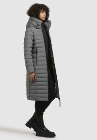 Gray padded coat with a high collar, zip closure, side pockets, and a long silhouette. Worn over black attire and black ankle boots.