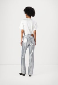 Pantaloni a palazzo metallici argentati con vita alta, due tasche posteriori e una toppa in pelle marrone, abbinati a una t-shirt bianca semplice.