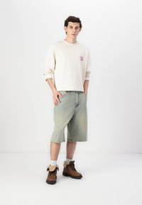 Camisa beige de manga larga con un pequeño gráfico, pantalones cortos de mezclilla azul claro y botas marrones con detalles en crema. Tejido de algodón, corte relajado.