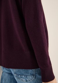 Burgunderroter Pullover mit weicher Textur, gerippten Bündchen und Saum, mit lockerem Schnitt. Kombiniert mit hellblauen Denim-Jeans.