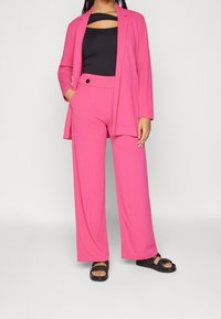 Blazer rosa y pantalones wide-leg a juego en una tela suave. Top negro con un diseño recortado. Combinado con sandalias negras de tipo slide.