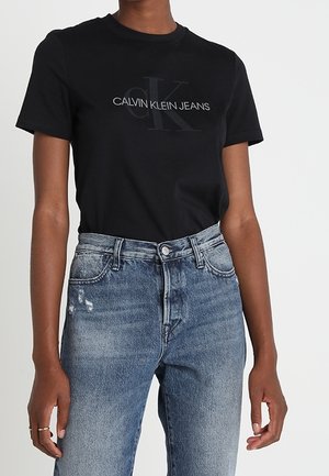 Czarny bawełniany t-shirt z wypukłym ciemnym logo "CALVIN KLEIN JEANS", w zestawieniu z jasnoniebieskimi, postrzępionymi dżinsami.