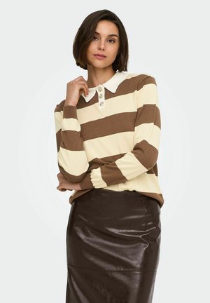 Femme aux cheveux bruns courts portant un pull à rayures marron et crème avec un col blanc et une jupe en cuir marron foncé.