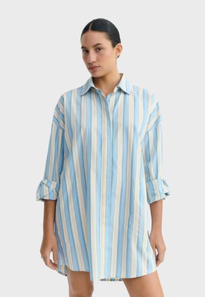 Kobieta o ciemnych włosach nosi luźną, długą sukienkę koszulową w niebiesko-białe pionowe paski z podwiniętymi rękawami, stojąc na jednolitym tle.