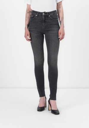 Persoon die een hooggetailleerde vervaagde zwarte skinny jeans, zwarte hoge hakken, een witte cropped top en zichtbare armtattoeages draagt.