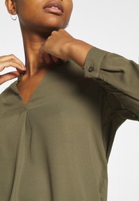 Oliwkowa bluzka z dekoltem w serek i długimi rękawami, uszyta z gładkiego materiału; posiada subtelną plisę przy dekolcie oraz mankiet zapinany na guziki.