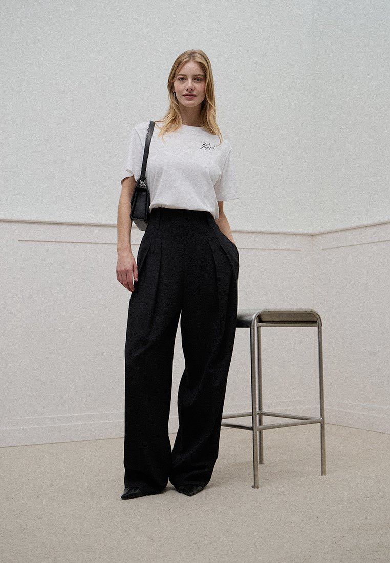 Weißes Baumwoll-T-Shirt mit schwarzem Logo, kombiniert mit hoch taillierten schwarzen weit geschnittenen Hosen. Das Model hält eine kleine schwarze Tasche und steht auf einem beigen Boden.