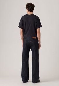 Man die met zijn rug naar de camera staat, gekleed in een eenvoudig zwart T-shirt, zwarte wijde jeans en zwarte schoenen tegen een effen achtergrond.