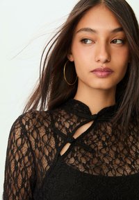 Sort blonde top med høj krave, nøglehulssnit og dekorative knuder. Designet har et blomstermotiv og en tekstureret overflade.
