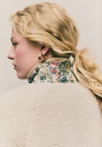 Kræmfuglet fleecejakke med en broderet blomsterkrave, der har pastelfarver. Personen bærer en spiralformet guldørering.
