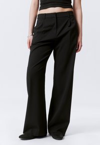 Woman wearing high-waisted black wide-leg trousers, black top, black shoes, and a silver bracelet against a plain light background.