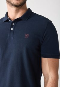 Camisa polo azul marino con un tejido texturizado, tapeta de dos botones y un pequeño logo burdeos en el pecho. Mangas cortas con puños acanalados.