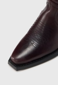 Σκούρο καφέ δερμάτινη μπότα με ραμμένα διακοσμητικά σχέδια και τετράγωνη μύτη σε μαύρη σόλα, σε λευκό φόντο.