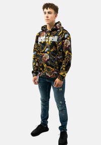 Felpa nera con cappuccio, decorata con un motivo barocco colorato e testo bianco. Abbinata a jeans blu strappati e scarpe da ginnastica nere.