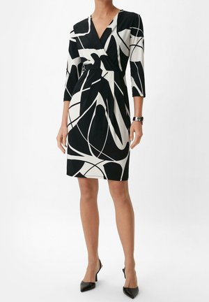Kvinde iført knælængde kjole med sort og hvidt abstrakt print, trekvartærmer og sorte spidse højhælede sko, stående foran ensfarvet baggrund.