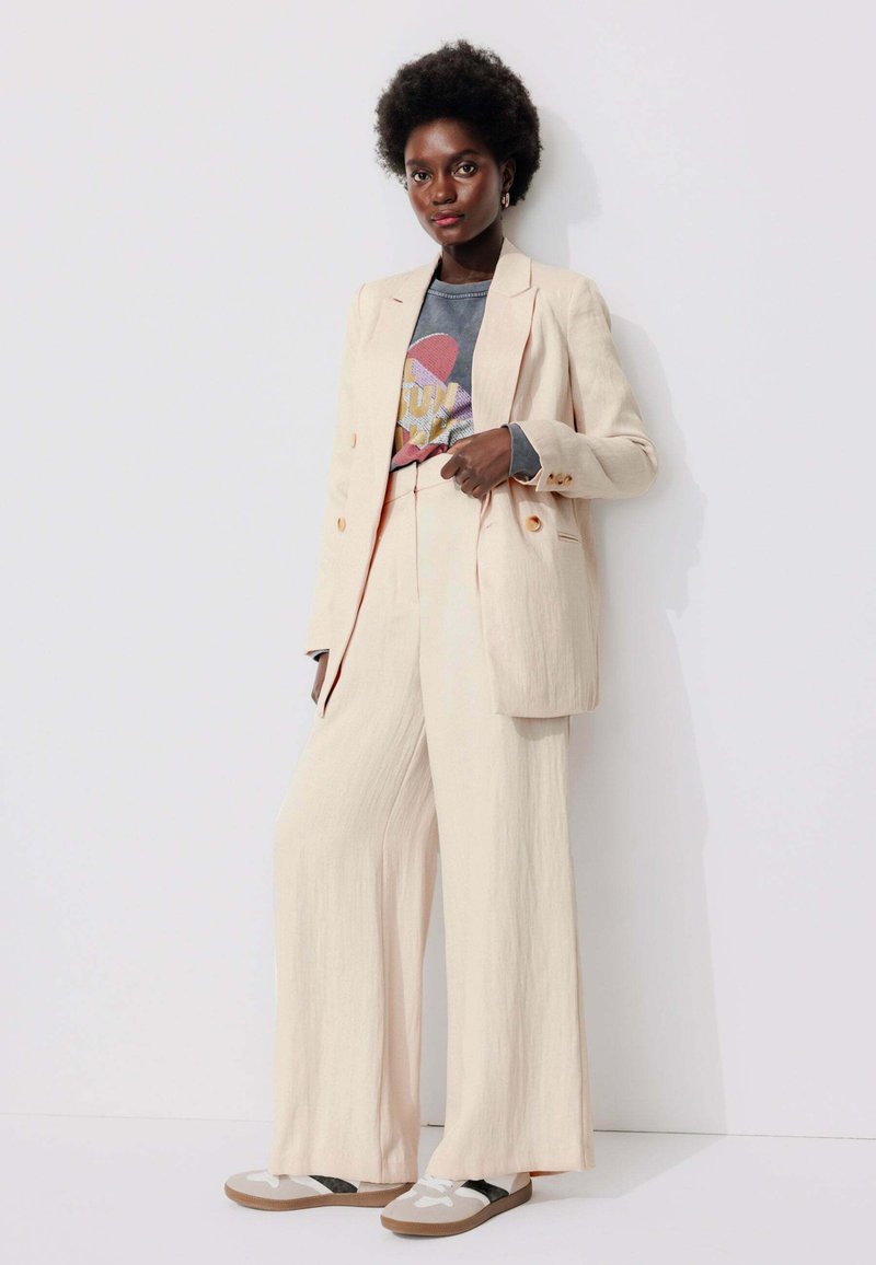 Costume en lin beige avec un pantalon à jambes larges et un blazer croisé sur un t-shirt graphique. Assorti avec des baskets décontractées.