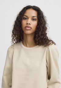 Beiger Pullover mit weicher Textur, rundem Ausschnitt und Puffärmeln. Minimalistisches Design ohne Muster oder Verzierungen.
