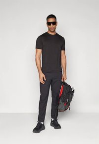 Svart t-shirt och svarta byxor, båda åtsittande. Svart ryggsäck med röda accenter hålls i en hand. Svarta sneakers och mörka solglasögon fulländar looken.