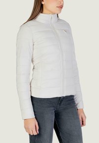 Giaccone imbottito bianco con collo alto, motivo trapuntato verticale, cerniera frontale e tasche laterali, abbinato a jeans scuri. Materiale liscio e leggero.