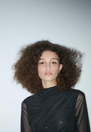 Zwarte doorschijnende blouse met een hoge hals, versierd met kleine sprankelende details, gecombineerd met volumineus krullend haar en zilveren oorbellen.