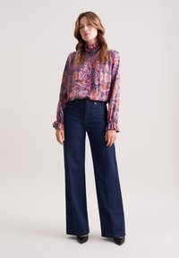 Bunte Paisley-Print-Bluse mit Rüschensaum, kombiniert mit hochgeschnittenen, dunkelblauen weitläufigen Jeans im klassischen Fünf-Taschen-Design.