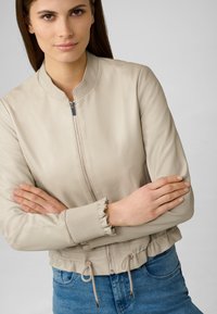 Beige Leder Bomberjacke mit Stehkragen, Reißverschluss vorne, gerippten Bündchen und verstellbarem Saum, kombiniert mit blauen Jeans.