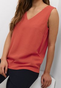Coral sleeveless top with a V-neck, made of a smooth fabric; features a subtle pocket detail and a relaxed fit, paired with dark pants.