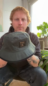 Zwarte emmerhoed van gestructureerde stof met een voorlabel met de tekst "Barbour", een afgeronde kroon en een brede, flexibele rand.