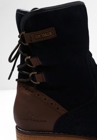 Botte en daim bleu foncé avec des accents en cuir marron, lacets décoratifs et détails perforés. Comprend une étiquette de marque sur le côté.