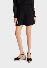 Pantaloni corti neri con pieghe, abbinati a sandali con zeppa neri e grigi con punta aperta, cinturino alla caviglia e suola intrecciata.