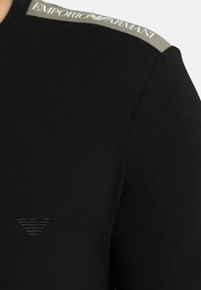 T-shirt noir en coton, avec un logo embossé discret sur la poitrine et une étiquette de marque grise contrastante sur l'épaule.