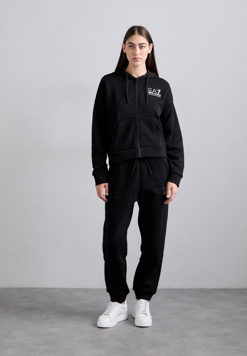 Woman stands against gray wall wearing black EA7 hooded zip sweatshirt, matching sweatpants, and white sneakers, facing forward with arms relaxed.