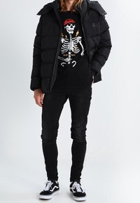 Giacca imbottita nera sopra una maglietta nera con una grafica di scheletro colorata, abbinata a jeans skinny neri e sneakers nere.