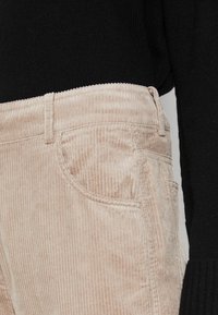 Pantalon en velours côtelé souple de couleur beige clair avec des côtes verticales, doté de poches standards et d'une ceinture ajustée, associé à un pull en maille noir.