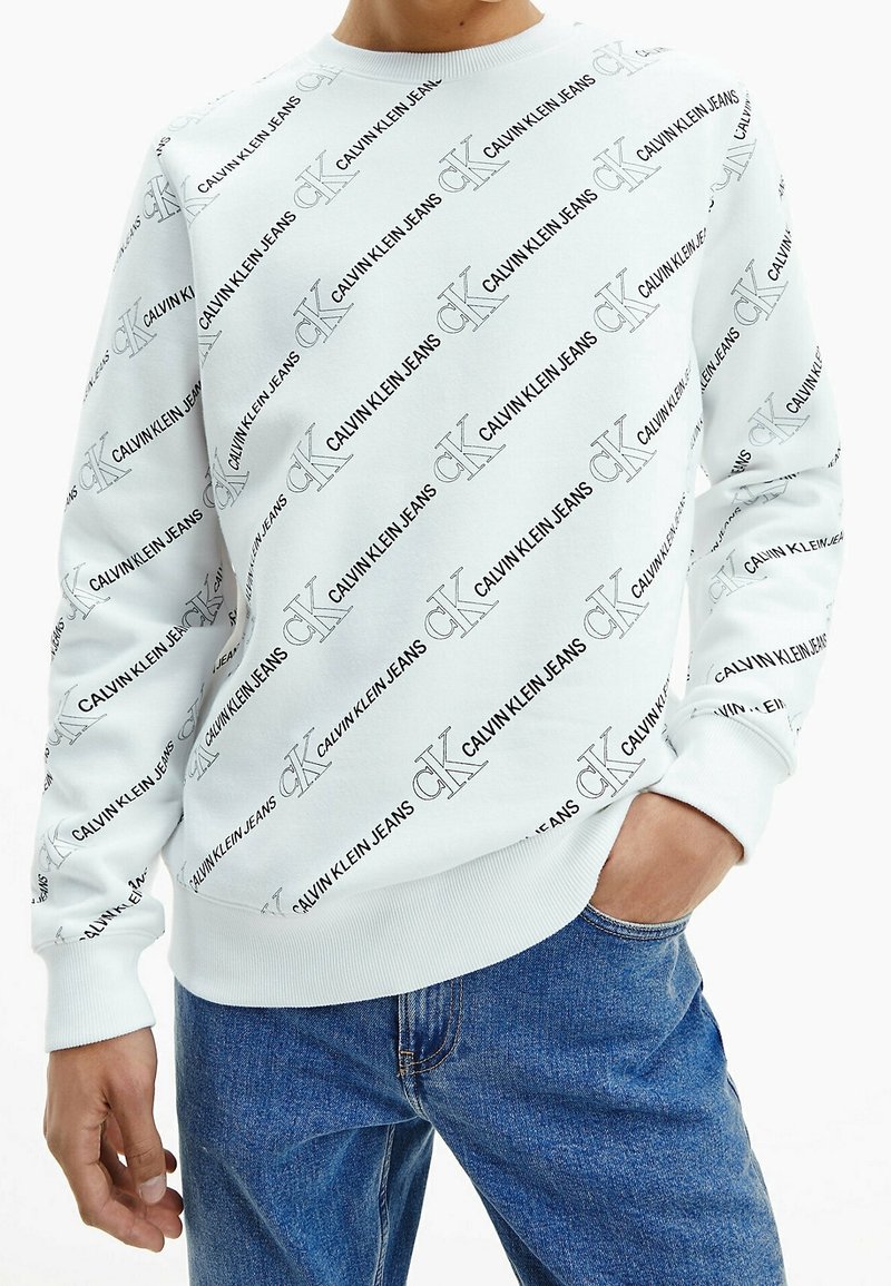 Felpa bianca con motivo logo "Calvin Klein Jeans" in nero su tutta la superficie, scollatura rotonda, polsini e orlo a coste; abbinata a jeans in denim blu.