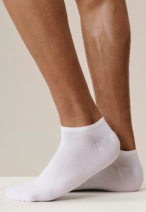 Para brązowych nóg w gładkich, białych skarpetkach sięgających za kostkę, z jedną stopą lekko uniesioną na neutralnym tle.