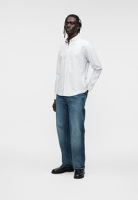 Λευκό πουκάμισο με κουμπιά, μπλε τζιν, μαύρα παπούτσια. Το πουκάμισο έχει κλασικό γιακά· το τζιν έχει άνετη γραμμή και ελαφρώς ξεθωριασμένο χρώμα.