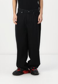 Black wide-leg jeans made of denim, featuring a button closure, belt loops, and front pockets. Worn with black and red sporty shoes.