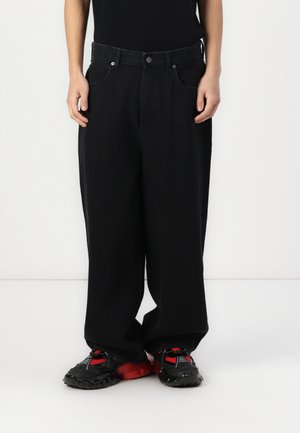 Jean large noir en denim, avec une fermeture à bouton, des passants de ceinture et des poches avant. Porté avec des chaussures de sport noires et rouges.