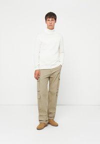 Joven de pie en pantalones cargo beige, suéter de cuello alto blanco y zapatillas color beige frente a un fondo blanco liso.