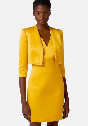 Donna che indossa un vestito aderente giallo acceso con una giacca corta abbinata con lettere ricamate sulla parte anteriore.