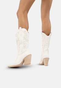 Botas de cowboy blancas con puntera puntiaguda, caña curvada y un intrincado bordado tonal. El tacón es de un tono claro de madera.