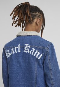 Jeans jakna s krznim ovratnikom, na hrbtu s velikim belim napisom "Karl Kani". Temno modra tkanina, strukturiran kroj, vidno šivanje.